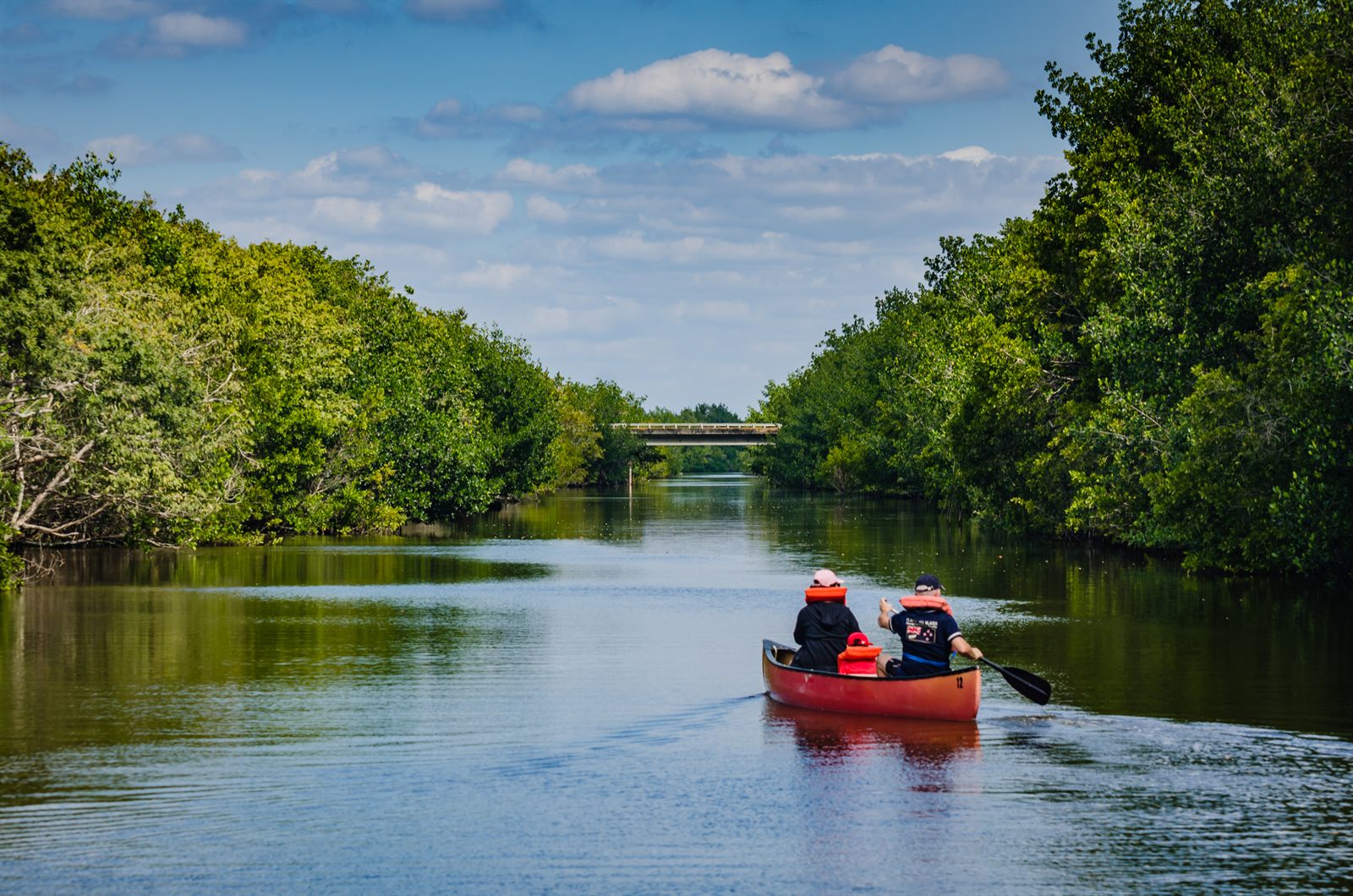 Two people paddling a red canoe on a calm river between green trees, with a bridge in the distance